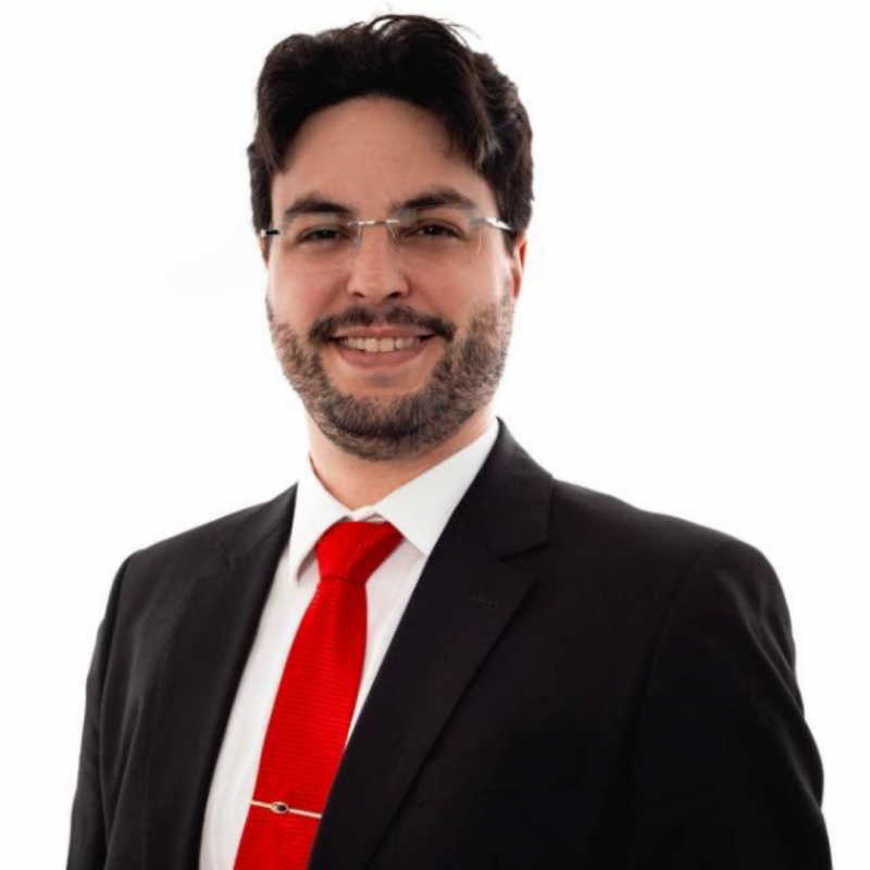 A imagem mostra um homem de terno sobre um fundo branco. O homem possui cabelo escuro curto, pele clara, barba aparada. Ele veste um terno preto com camisa branca e gravata vermelha. Ele usa óculos.
