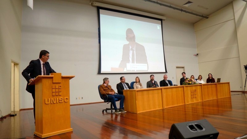 A foto mostra um palco com uma mesa e um púlpito. Atrás da mesa, estão sentadas 9 pessoas, enquanto no púlpito um homem discursa.