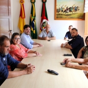 Em traje formal, os representantes do Procon RS e do Procon Municipal de Piratini estão sentados ao redor de uma mesa comprida e retangular. Ao fundo, estão hasteadas as bandeiras do Brasil, Rio Grande do Sul e do município de Piratini.