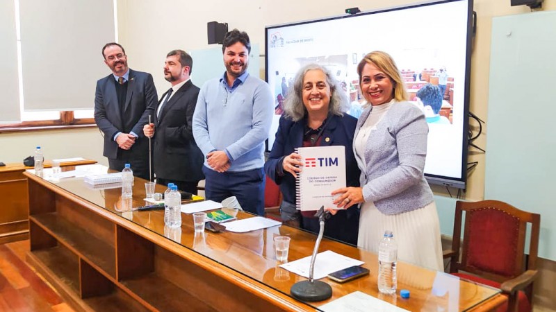 Autoridades posicionadas na mesa de abertura, uma mesa de madeira coberta por um tampão de vidro. As autoridades seguram o documento em braille.