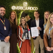Seis pessoas estão posando para uma foto em um ambiente interno, diante de uma parede verde de plantas artificiais com o letreiro iluminado “@VERBOJURIDICO”. No centro, uma mulher de cabelos loiros e lisos veste blazer listrado em vermelho, azul e branco e segura uma revista com a capa exibindo sua própria foto. Ao lado dela, um homem de blazer azul e camisa branca segura um documento impresso. Os demais, três mulheres e um homem, estão sorridentes, vestidos com roupas sociais em tons de marrom, azul e bege, compondo uma atmosfera formal e cordial de registro institucional.